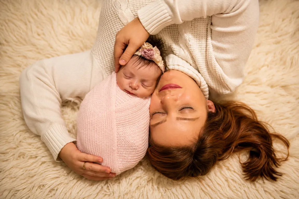 Rebeca resting cheek to cheek with a sleeping newborn on a soft cream blanket